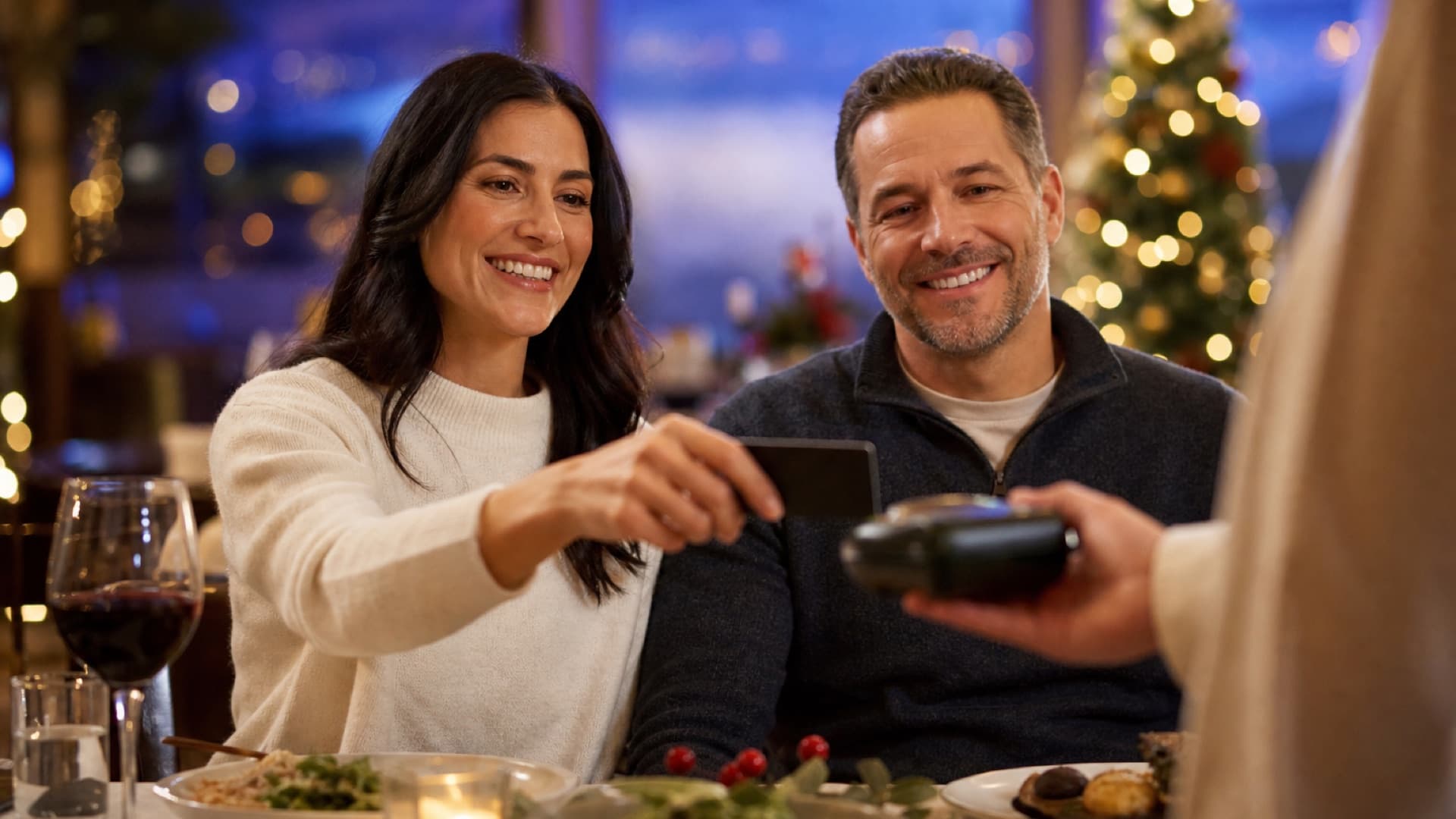 A smiling couple pays the bill for a holiday meal at a restaurant with a credit card, in front of a lit Christmas tree