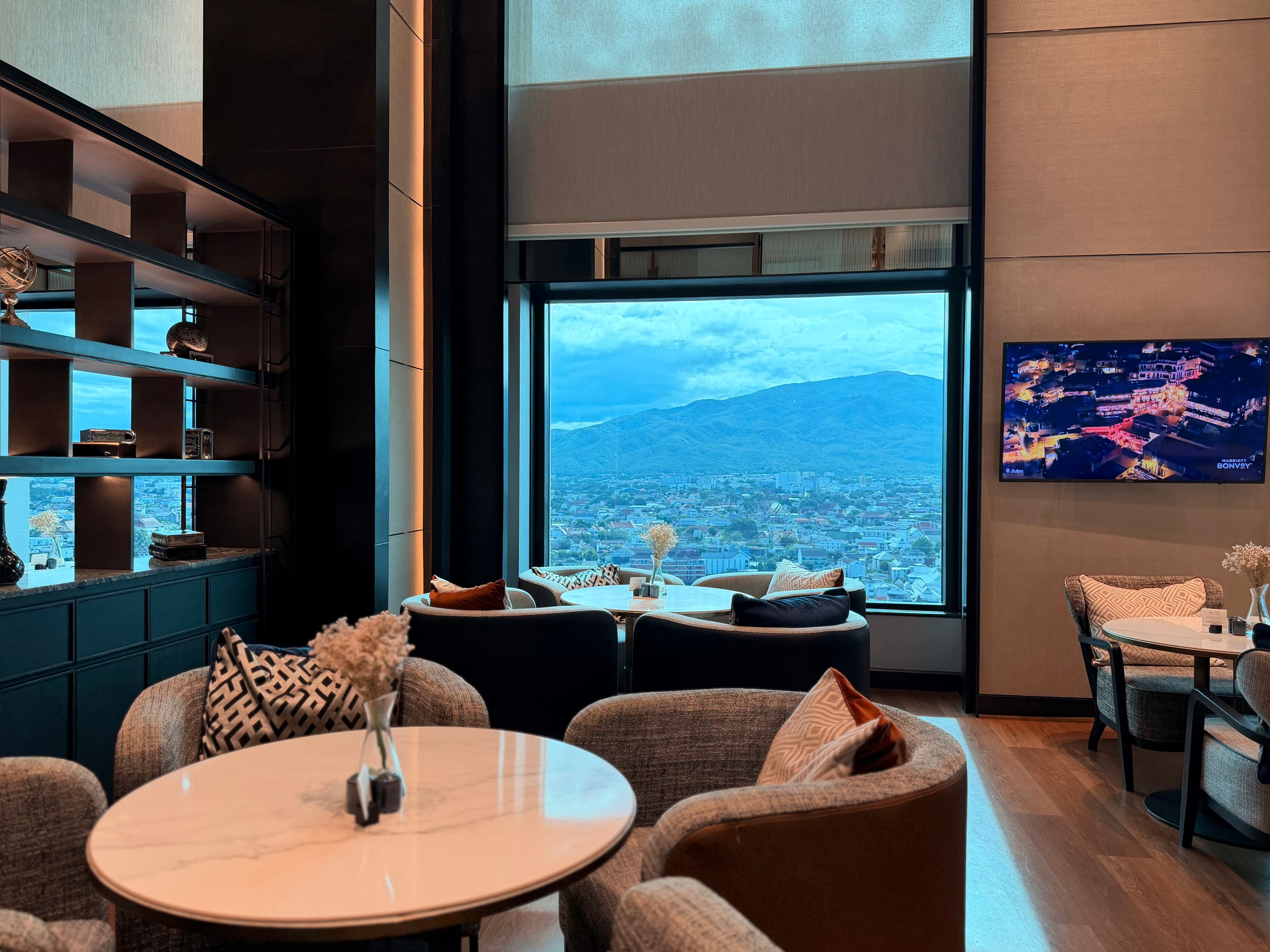 Espace avec tables rondes, grande baie vitrée sur la ville et écran Marriott Bonvoy au Marriott Chiang Mai