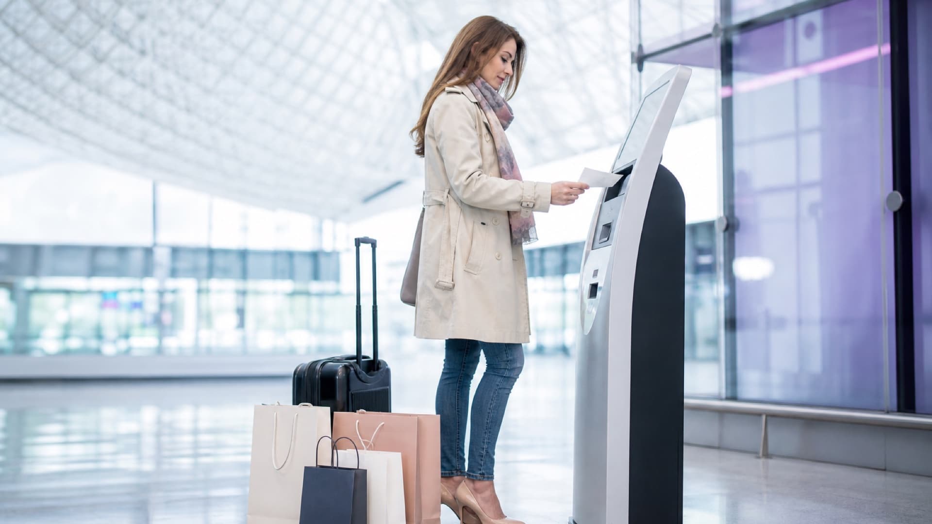 Traveller inserting a receipt into a tax-refund kiosk at a European airport, with shopping bags and a suitcase at her feet