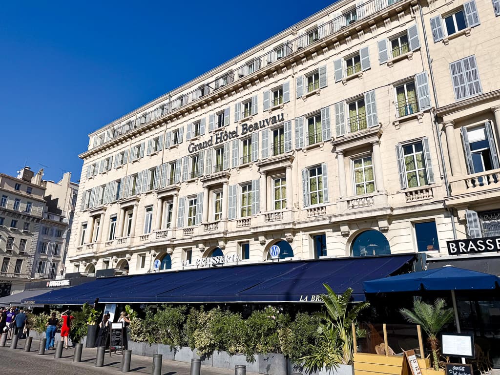 Façade blanche du Grand Hôtel Beauvau à Marseille, avec ses balcons en fer forgé et la terrasse de la brasserie sous un auvent bleu, sous un ciel bleu