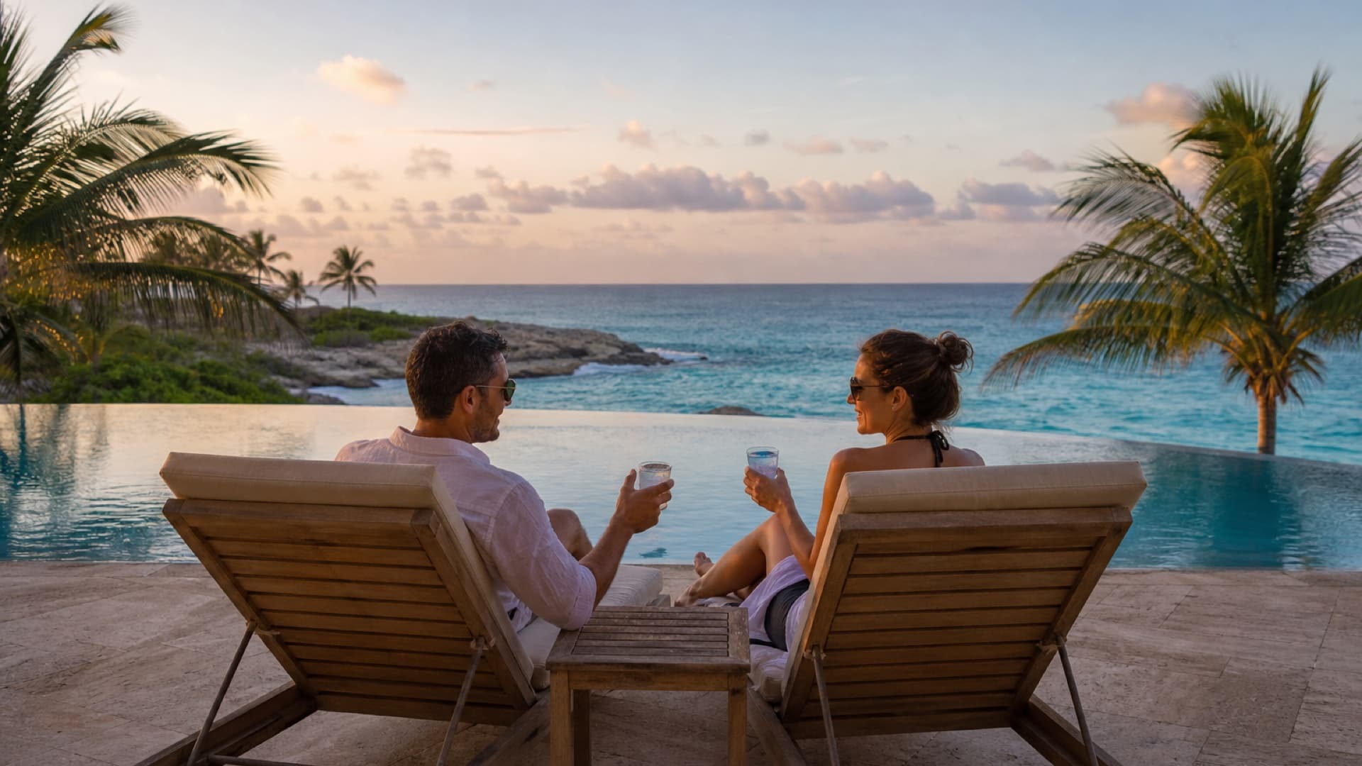 Couple sur des chaises longues au bord d'une piscine à débordement face à la mer des Caraïbes, au coucher du soleil
