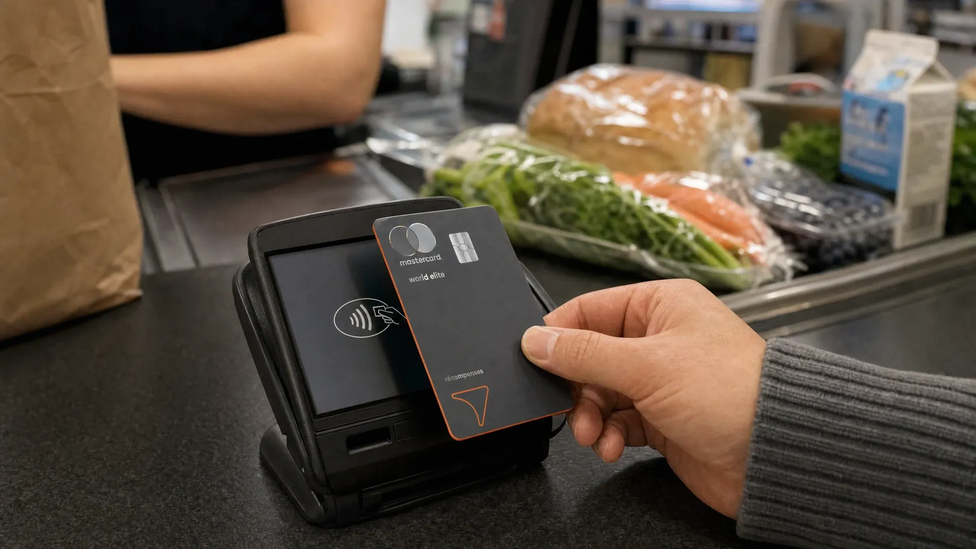 Une main tient la carte World Elite Mastercard Récompenses Tangerine près d'un terminal de paiement sans contact, à la caisse d'une épicerie