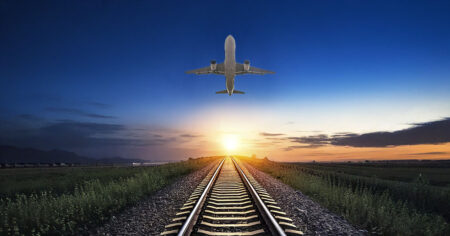 Un avion en vol au-dessus de voies ferrées, avec un coucher de soleil illuminant l'horizon