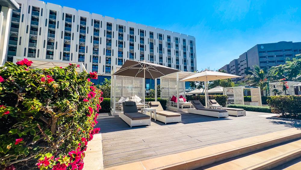 Pool deck du w muscat avec cabanes privatives blanches, parasols beiges, bougainvilliers roses et vue sur la façade moderne