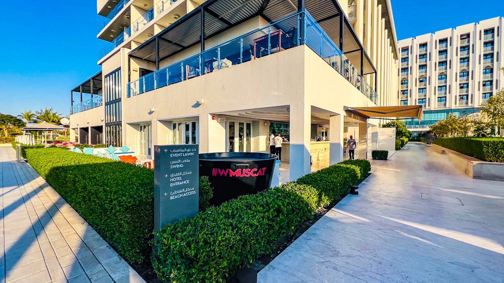 Entrée principale du w muscat avec architecture moderne beige, logo rose distinctif et jardins paysagers sous ciel bleu