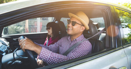 Conducteur souriant avec un chapeau et des lunettes de soleil, accompagné d'une passagère, dans une voiture.