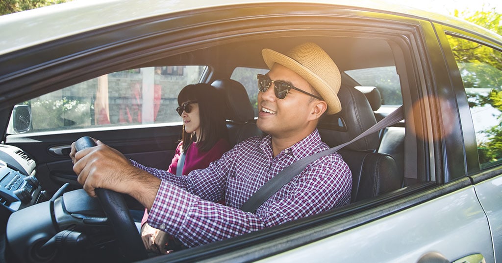 Conducteur souriant avec un chapeau et des lunettes de soleil, accompagné d'une passagère, dans une voiture.