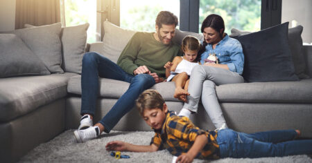 Famille souriante profitant d'un moment ensemble dans le salon. Les parents lisent avec leur fille sur le canapé tandis que le fils joue au sol avec ses jouets.