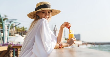 Une femme portant un chapeau de paille, savoure un jus d'orange en profitant d'une vue ensoleillée sur l'eau.