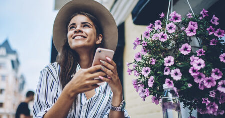 Une femme souriante, assise à l'extérieur, tient un téléphone et profite d'une ambiance ensoleillée près de fleurs violettes suspendues.