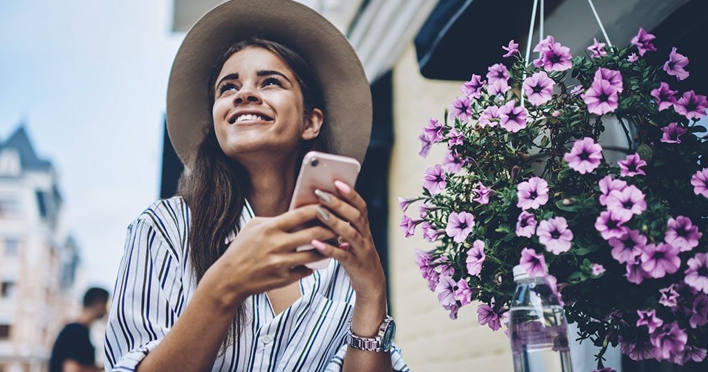 Une femme souriante, assise à l'extérieur, tient un téléphone et profite d'une ambiance ensoleillée près de fleurs violettes suspendues.