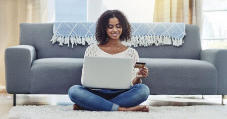 Une femme assise par terre utilise un ordinateur portable en tenant une carte de crédit, devant un canapé.