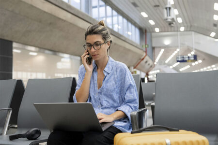 Une femme assise dans un aéroport utilisant un ordinateur portable et parlant au téléphone, avec des bagages à proximité