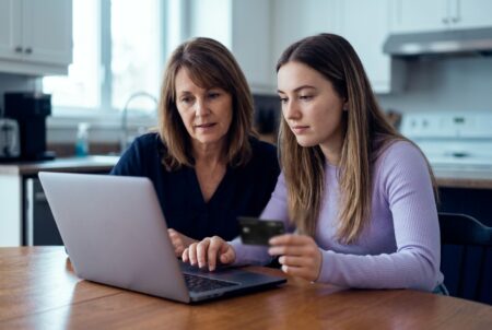 Une mère et sa fille adulte consultent un ordinateur portable en cuisine, la fille tenant une carte de crédit CIBC Dividendes Visa Infinite